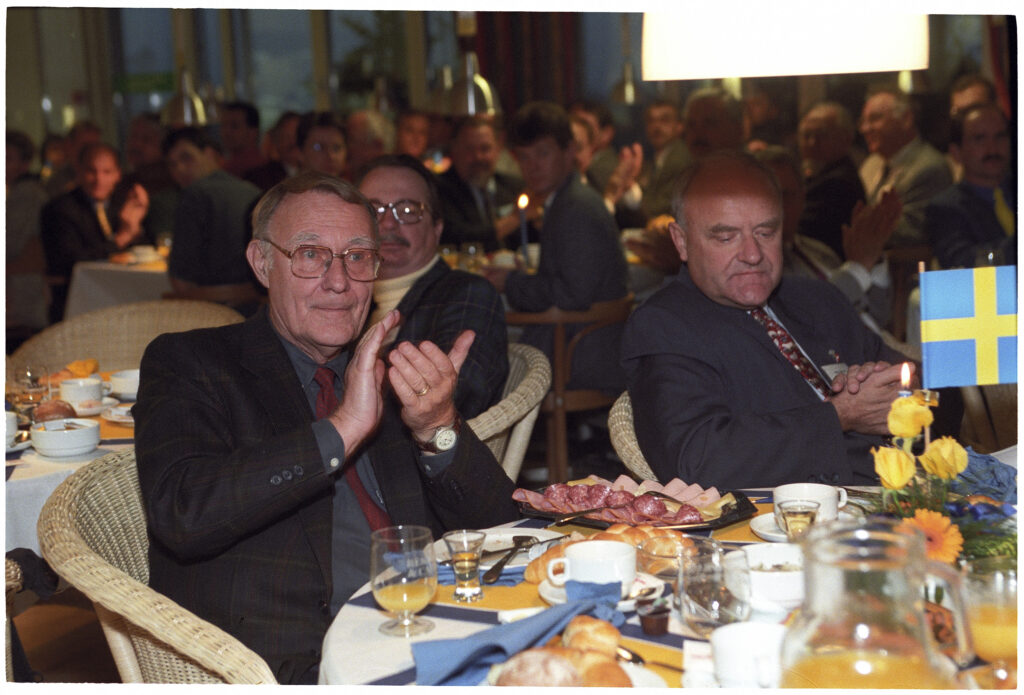 IKEA Gründer Ingvar Kampard sitzt mit einem weiteren Herren an einem Tisch und klatscht in die Hände. Vor ihm ein runder Tisch mit Gläsern, Kaffeetassen, Snacks und die schwedische Fahne
