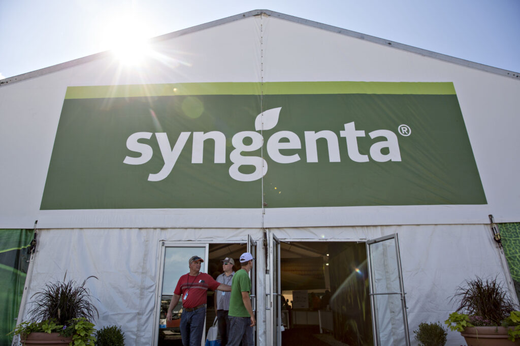 Logo von Syngenta in weiß und grün auf einer Halle montiert und von unten fotografiert. Vor der Halle stehen einige Menschen
