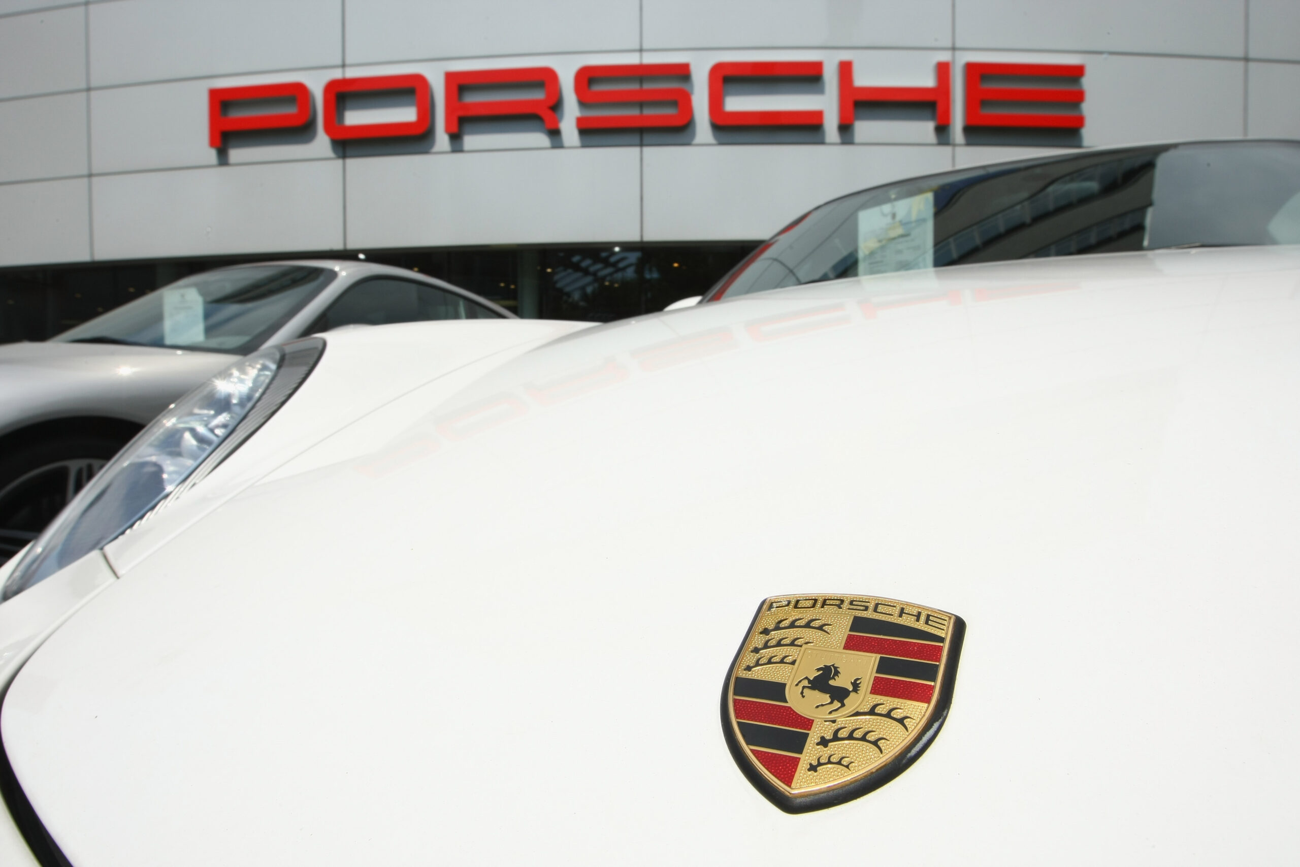 weiße Motorhaube mit Porsche-Logo und dahinter der Schriftzug PORSCHE in rot auf einem Gebäude