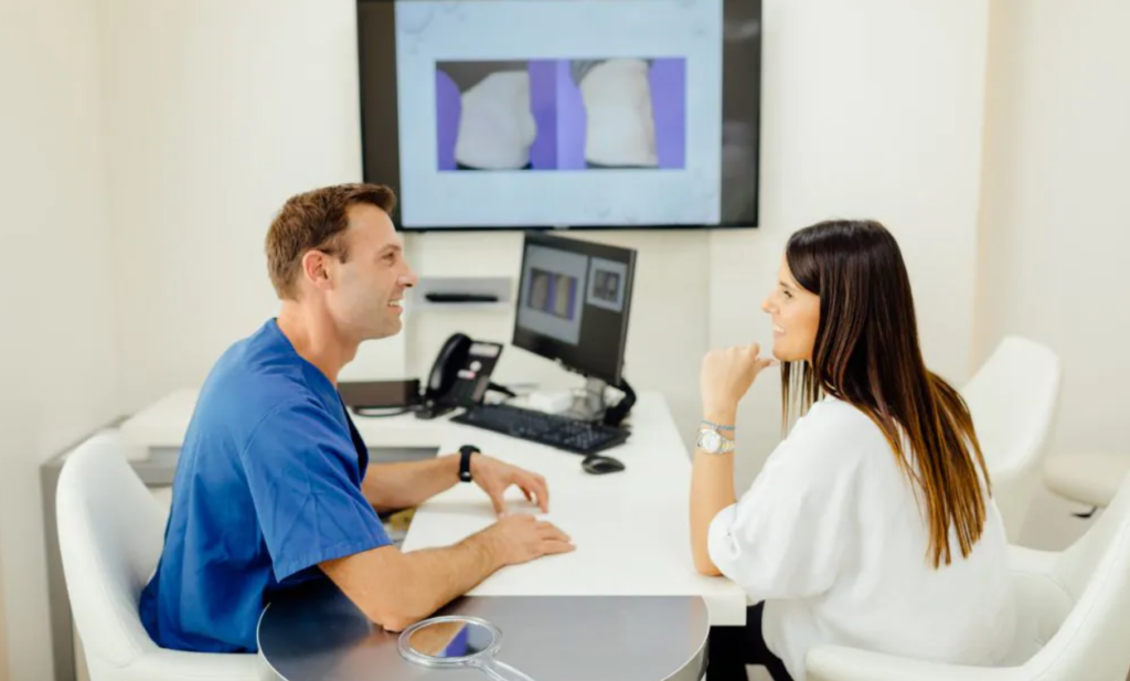 Schönheitschirurg René Draxler mit blauem Operationshemd sitzt einer Kundin im Beratungsgespräch über eine Brustvergrößerung gegenüber