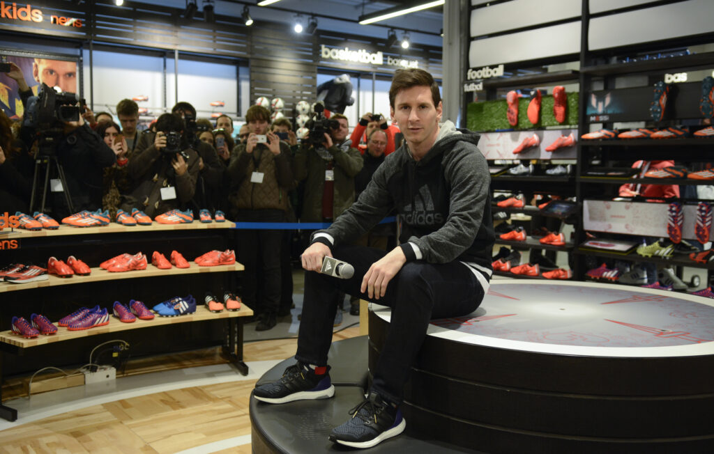 Lionel Messi sitzt mit einem Mikrofon in der Hand in einem Adidas-Store auf einem grunden Podium. Um ihn herum viele Schuhe und viele Reporter und Fans