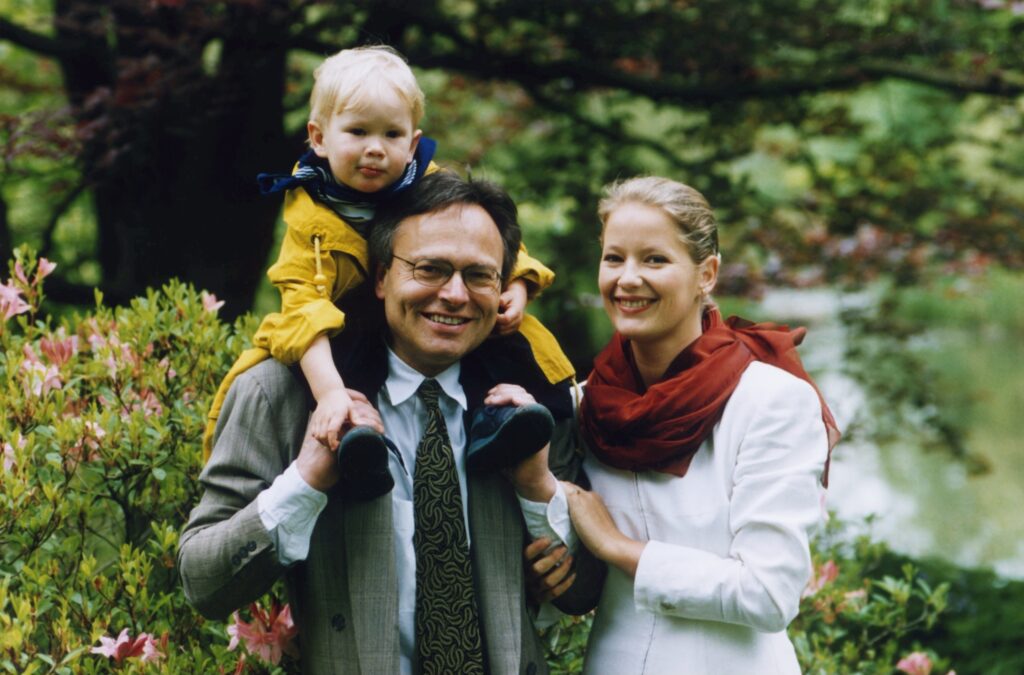 Günther Fielmann in jungen Jahren mit einem Kind auf seinen Schultern und seiner Frau mit weißer Bluse und rotem Schal neben ihm