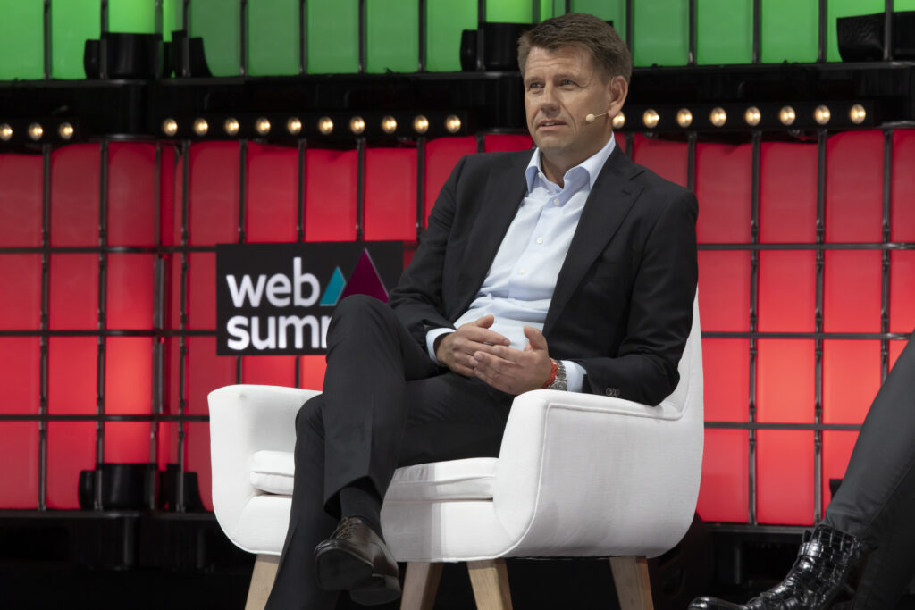 Oliver Steil als CEO von Teamviewer sitzt auf einer Bühne mit überschlagenen Beinen und hat ein Headset am Ohr. Hinter ihm ein roter und grüner Hintergrund mit websummit