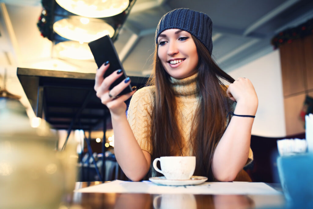 Frau mit Mütze und langen Haaren hält eine Handy in der Handy. Vor ihr steht eine Kaffeetasse auf ihrem Tisch