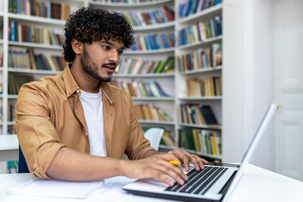 Junger Mann sitzt an einem PC und schreibt auf der Tastatur. Hinter im viele Bücher in hohen Regalen