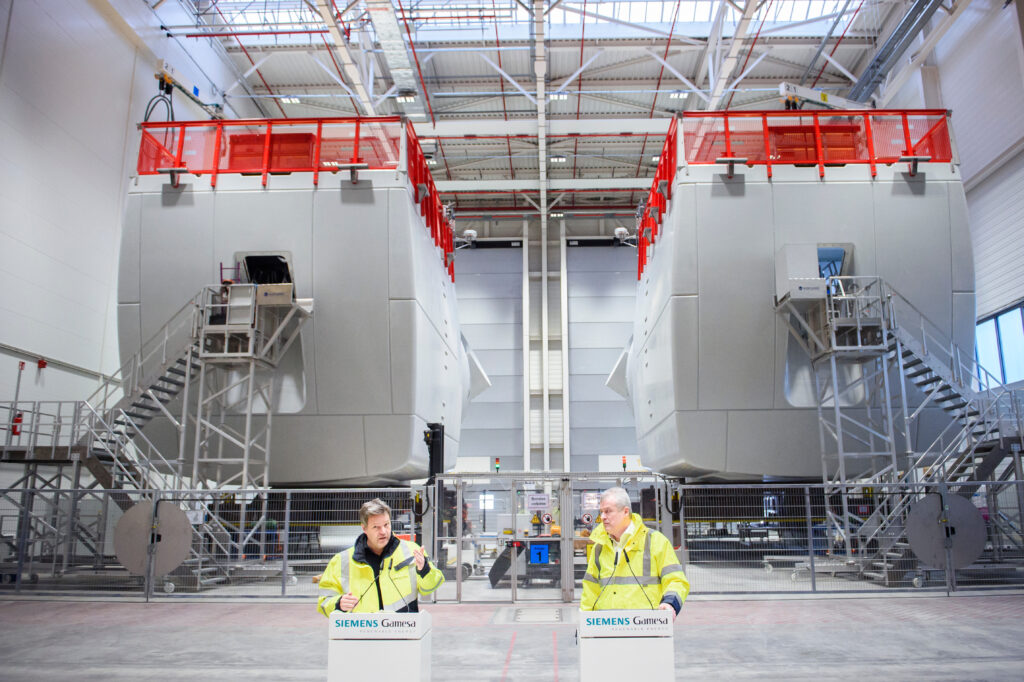 Blick in die Produktionshalle von Siemens Energy. Davor stehen zwei Herren ein einem Rednerpult. Beide tragen eine gelbe Jacke