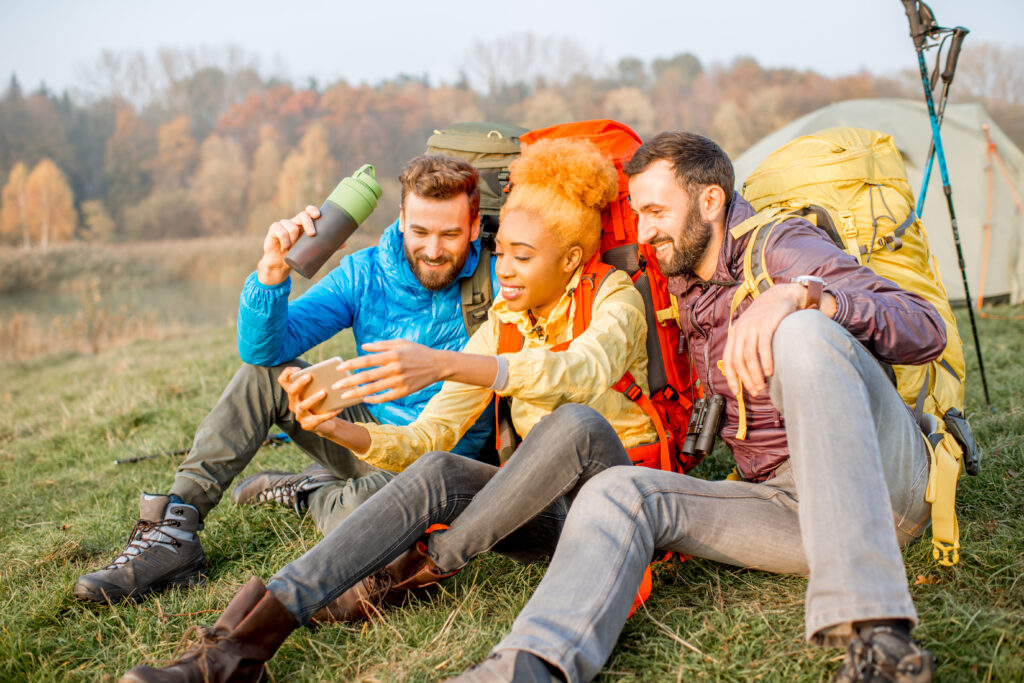 drei junge Menschen mit Rucksäcken sitzen auf einer Wiese und machen Selvies