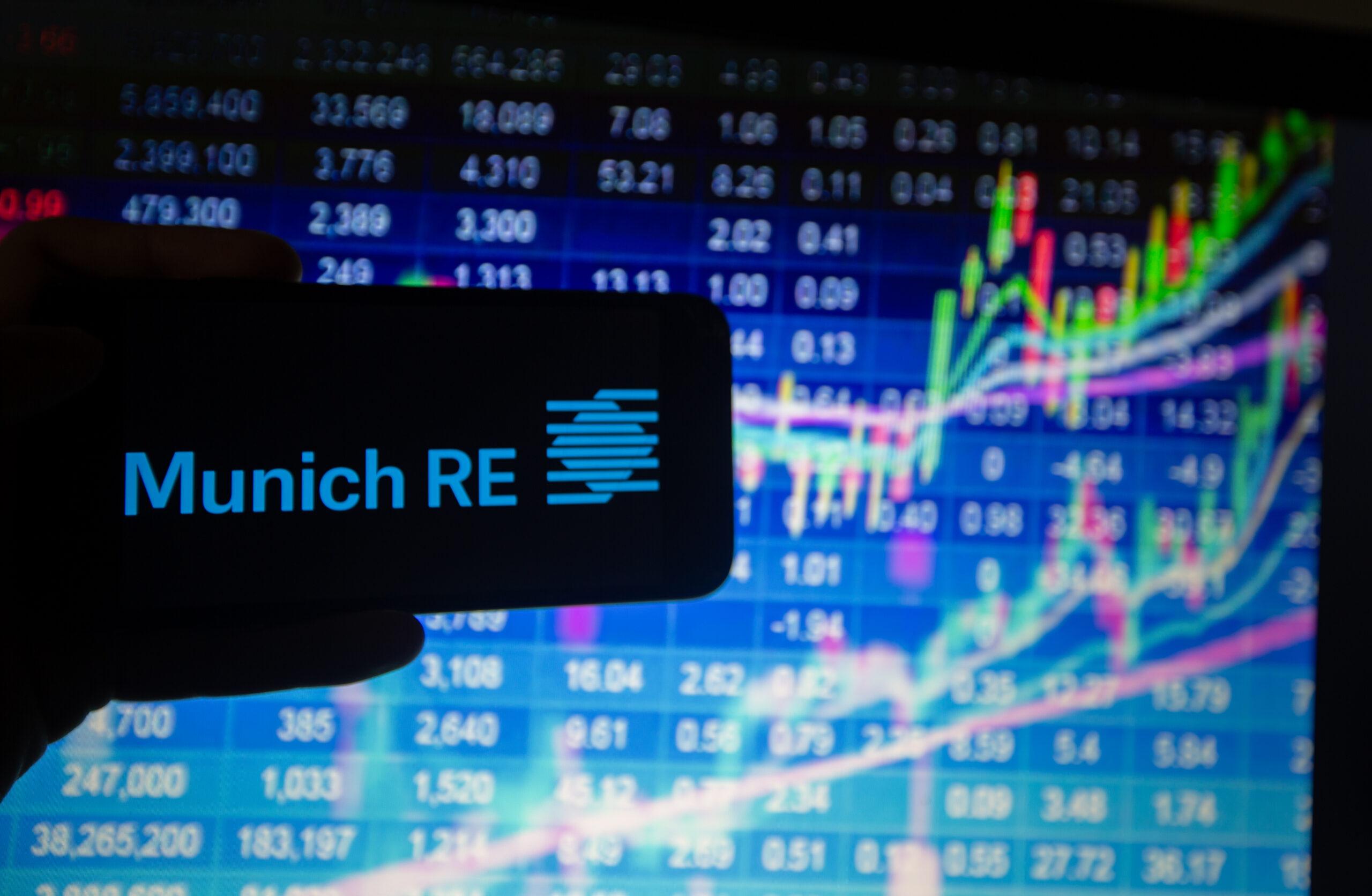 Logo von Munich Re auf einem Handy. Dahinter Börsenkurse